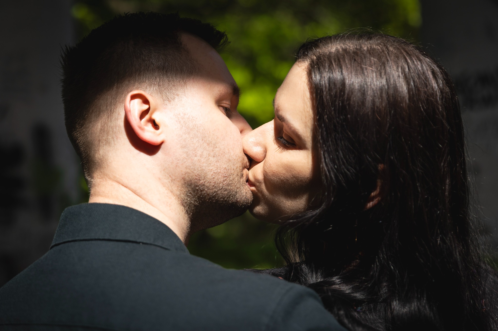 Zbliżenie na całującą się parę na tle zieleni. Mężczyzna w czarnej koszuli, kobieta z ciemnymi włosami. Oświetlenie słoneczne z cieniem.