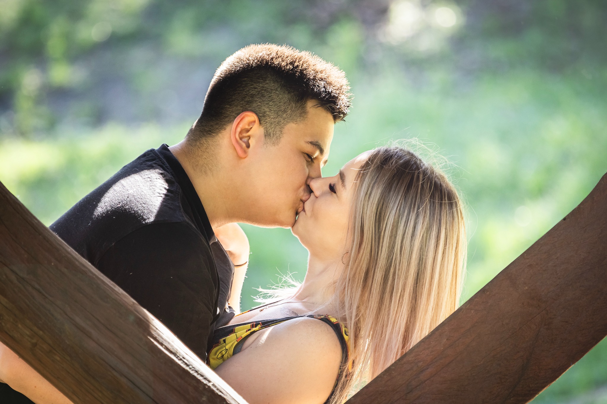 Zakochana para całuje się na tle zieleni, ujęcie zza drewnianej konstrukcji. Delikatne światło podkreśla intymność chwili. Romantyczna fotografia plenerowa.