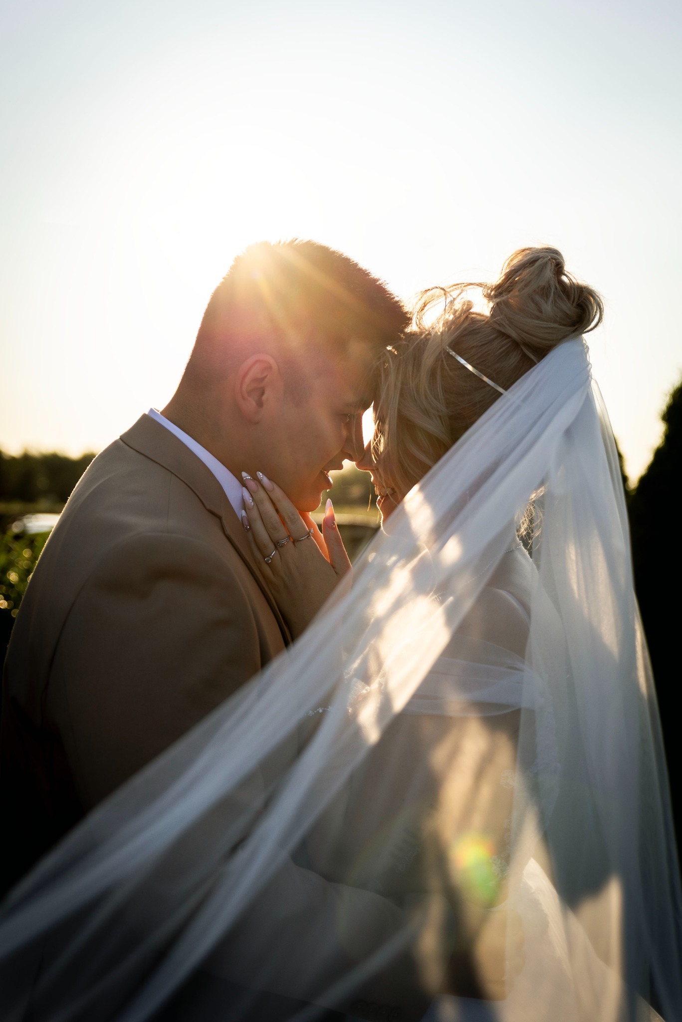 Para młoda w złocistym świetle zachodu, delikatny welon opada na twarz panny młodej, subtelne zbliżenie, romantyczna sesja ślubna w plenerze.