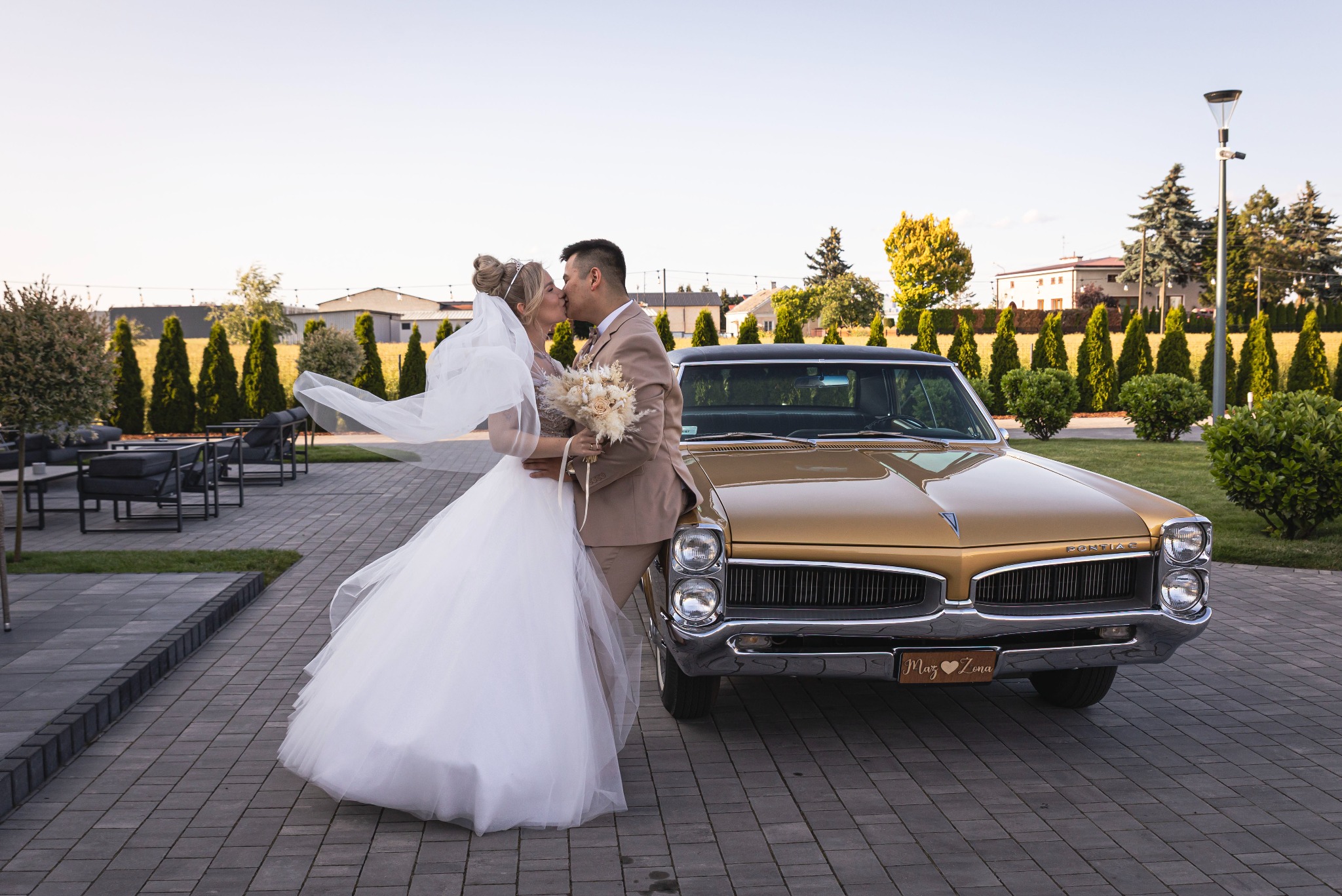 Para młoda w objęciach, całuje się na tle złotego, klasycznego Pontiaca z tablicą 'Mąż i Żona' w plenerze. W tle żywopłot i budynki.