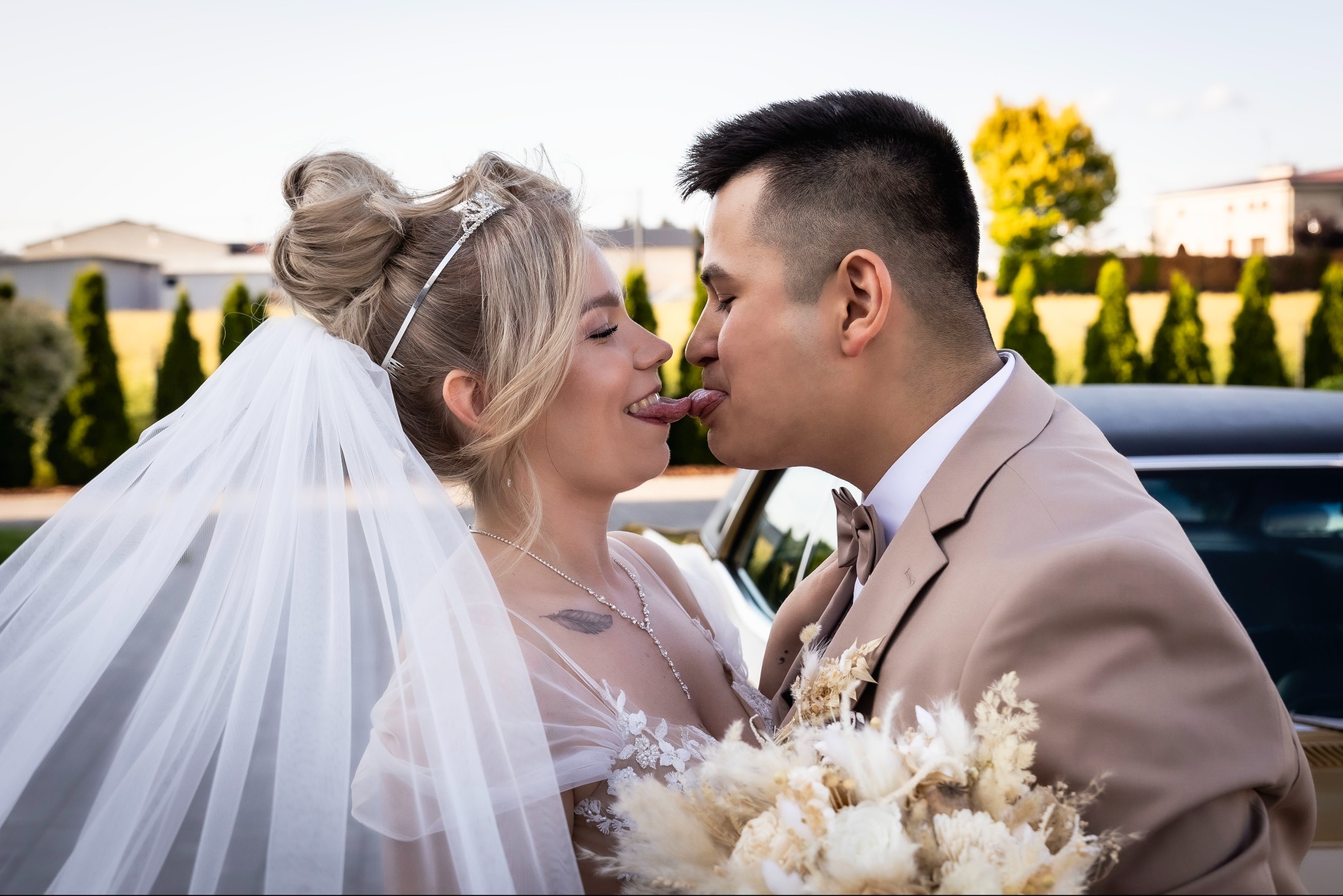 Nowożeńcy w beżowych strojach, stojący przed retro autem, wymieniają się językami. Panna młoda w tiarze i welonie, pan młody z bukietem. Nietypowe ujęcie.