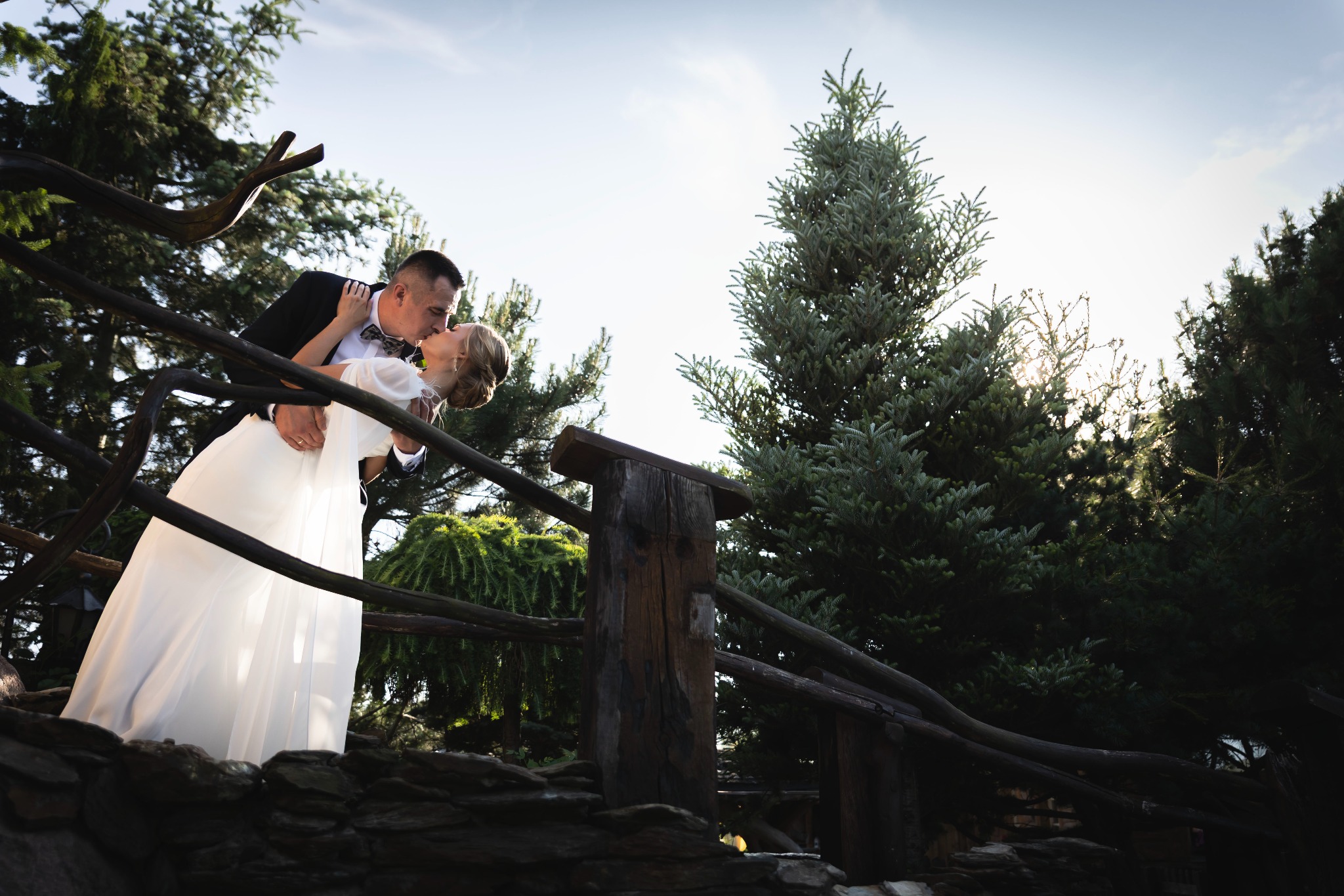 Pocałunek na drewnianym mostku: Para młoda w objęciach, ona w białej sukni, on w smokingu z muszką, otoczeni zielenią drzew. Romantyczna sceneria.