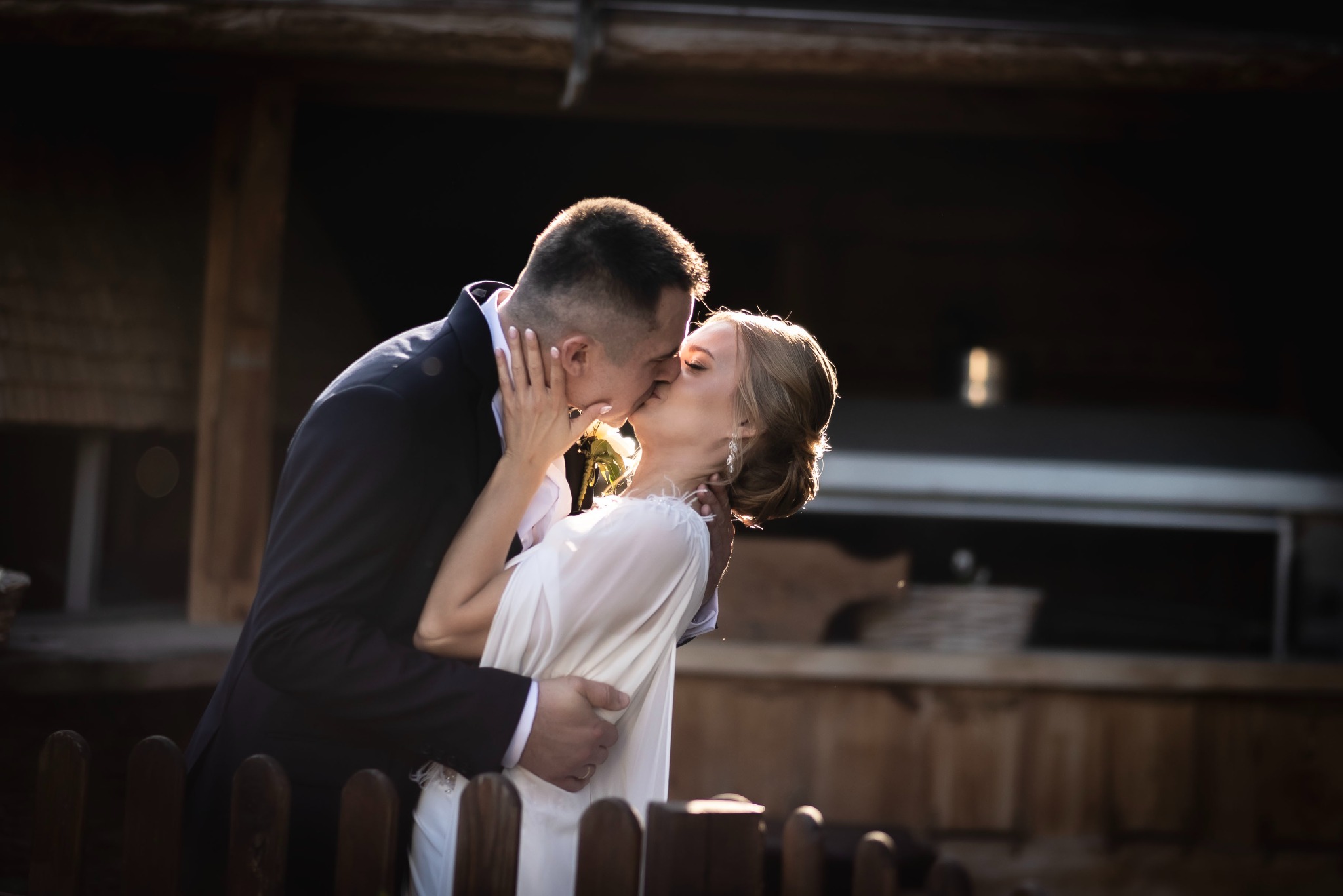 Para młoda w czułym pocałunku, złapana w kontrze do słońca. Delikatna suknia panny młodej i ciemny garnitur pana młodego na tle drewnianej konstrukcji.