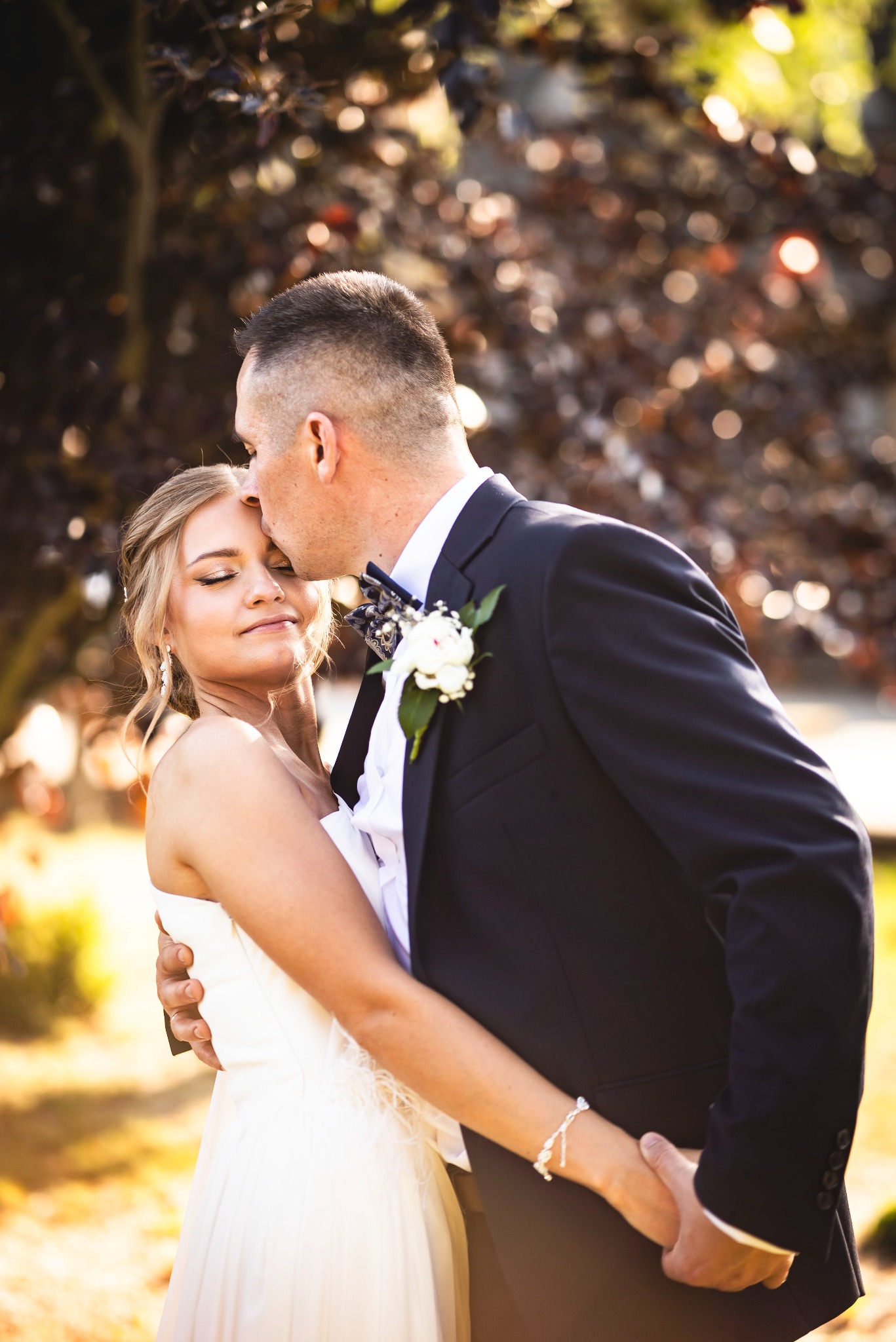 Elegancki pan młody całuje pannę młodą w czoło, trzymając ją w ramionach na tle rozmytej zieleni i ciepłych promieni słońca. Romantyczna chwila.