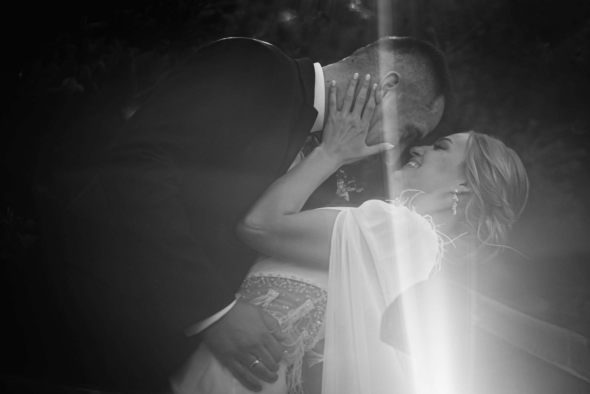 Czarno-białe zdjęcie pary młodej w objęciach, panna młoda uśmiechnięta, z rękami na twarzy pana młodego. Romantyczna scena z suknią ślubną i garniturem.