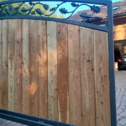 Drewniana brama wjazdowa z metalową ramą i ozdobnym motywem liści dębu w górnej części, widok z boku na podjazd i garaż.