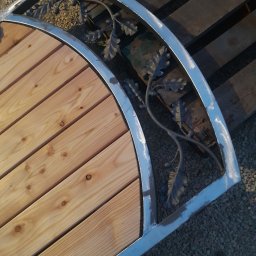 Stelmart Mariusz Lis - Fragment półokrągłej konstrukcji z jasnego drewna i metalu z dekoracyjnymi elementami liści dębu, leżącej na palecie i żwirze.