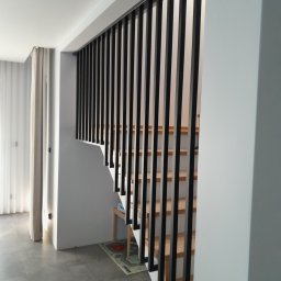 Stelmart Mariusz Lis - Nowoczesne schody z drewnianymi stopniami i czarną metalową balustradą w minimalistycznym wnętrzu z szarymi płytkami podłogowymi i białymi ścianami.