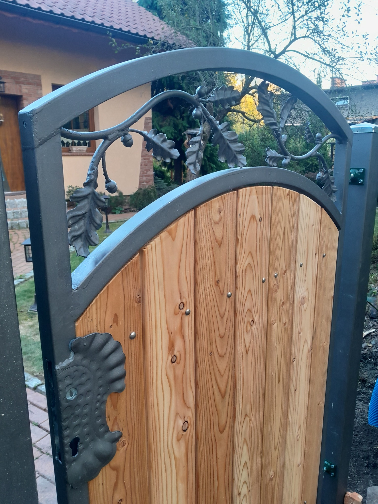 Drewniana furtka z metalową ramą i dekoracyjnymi elementami w kształcie liści dębu i żołędzi, z widocznym zamkiem w stylu retro.