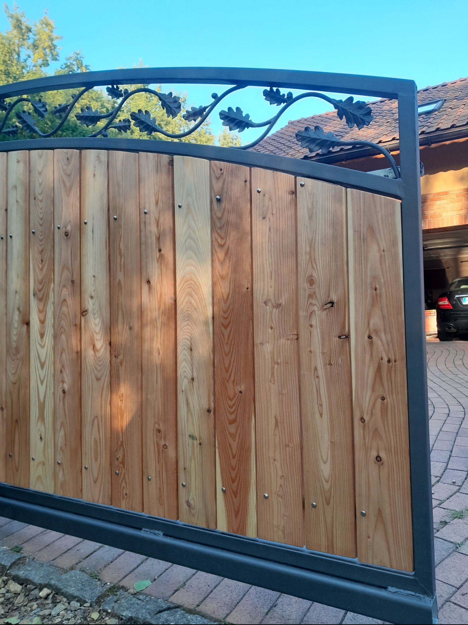 Drewniana brama wjazdowa z metalową ramą i ozdobnym motywem liści dębu w górnej części, widok z boku na podjazd i garaż.