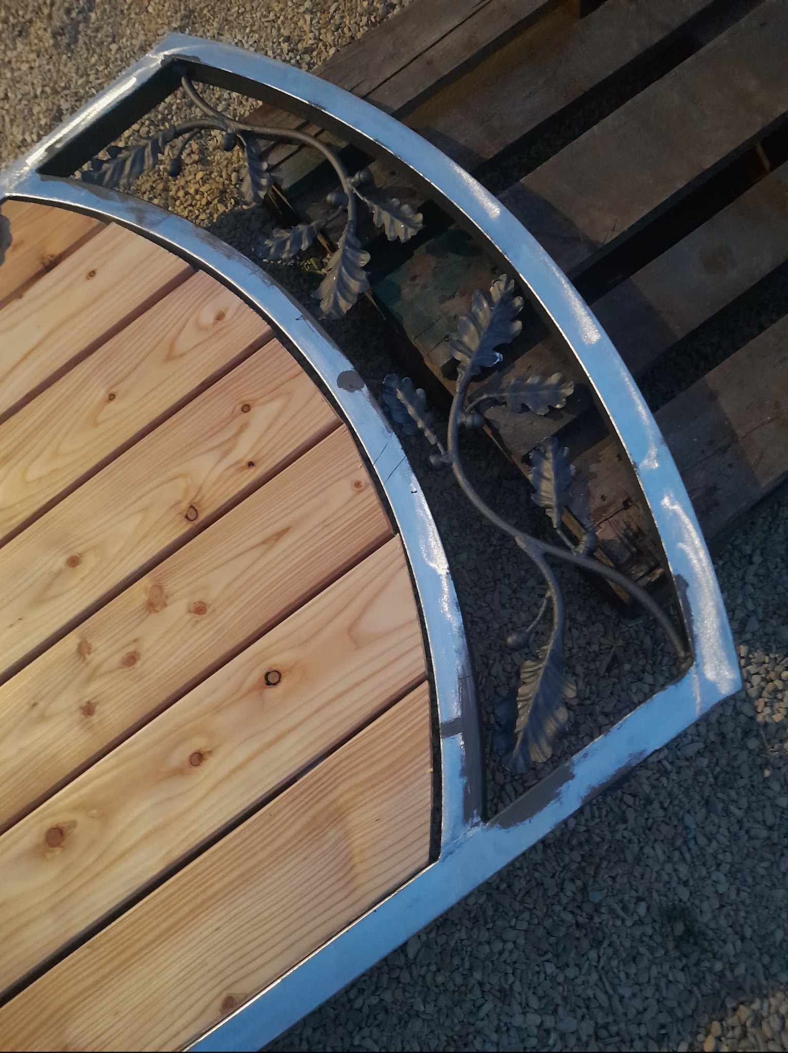 Fragment półokrągłej konstrukcji z jasnego drewna i metalu z dekoracyjnymi elementami liści dębu, leżącej na palecie i żwirze.