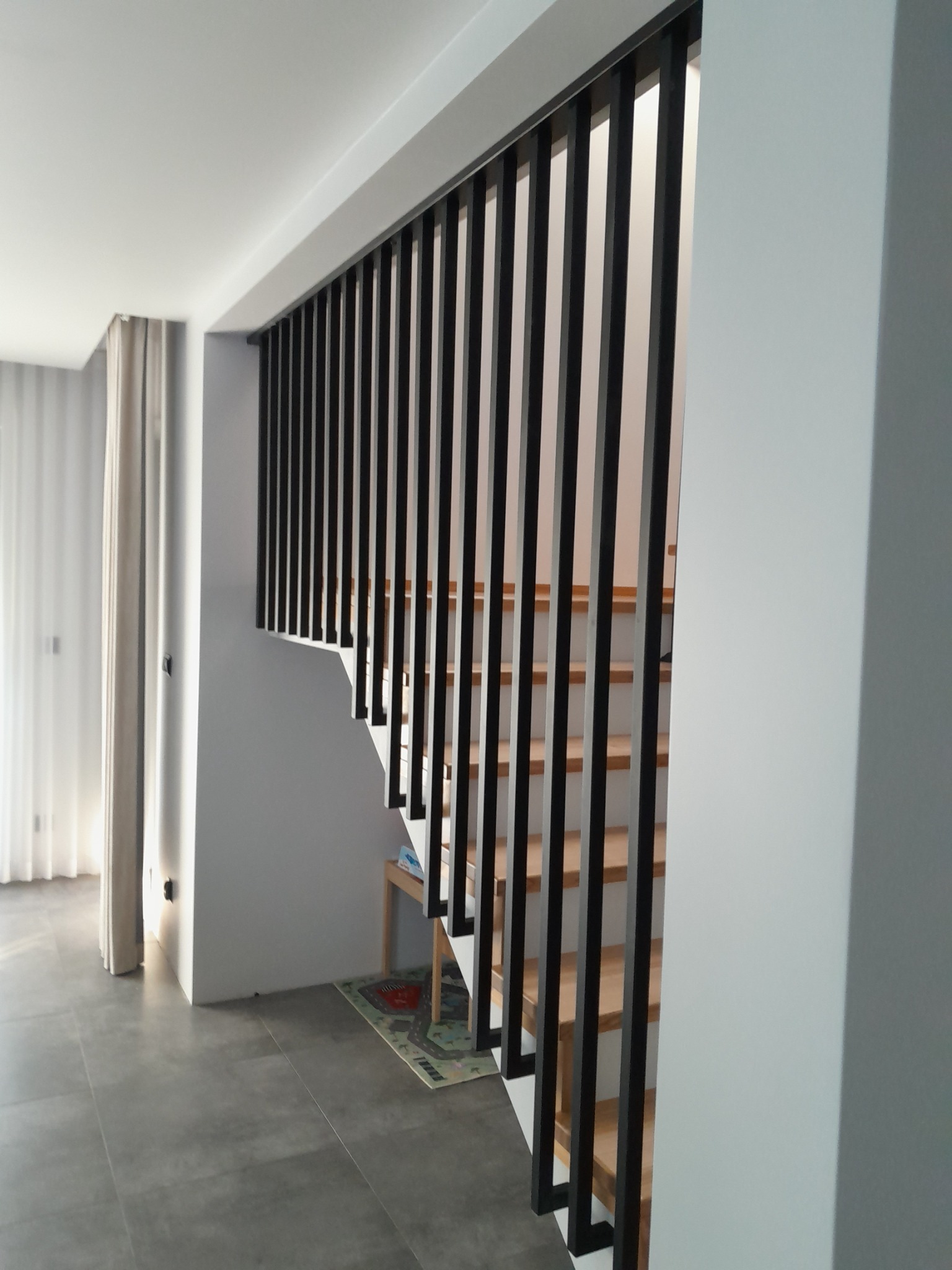 Nowoczesne schody z drewnianymi stopniami i czarną metalową balustradą w minimalistycznym wnętrzu z szarymi płytkami podłogowymi i białymi ścianami.