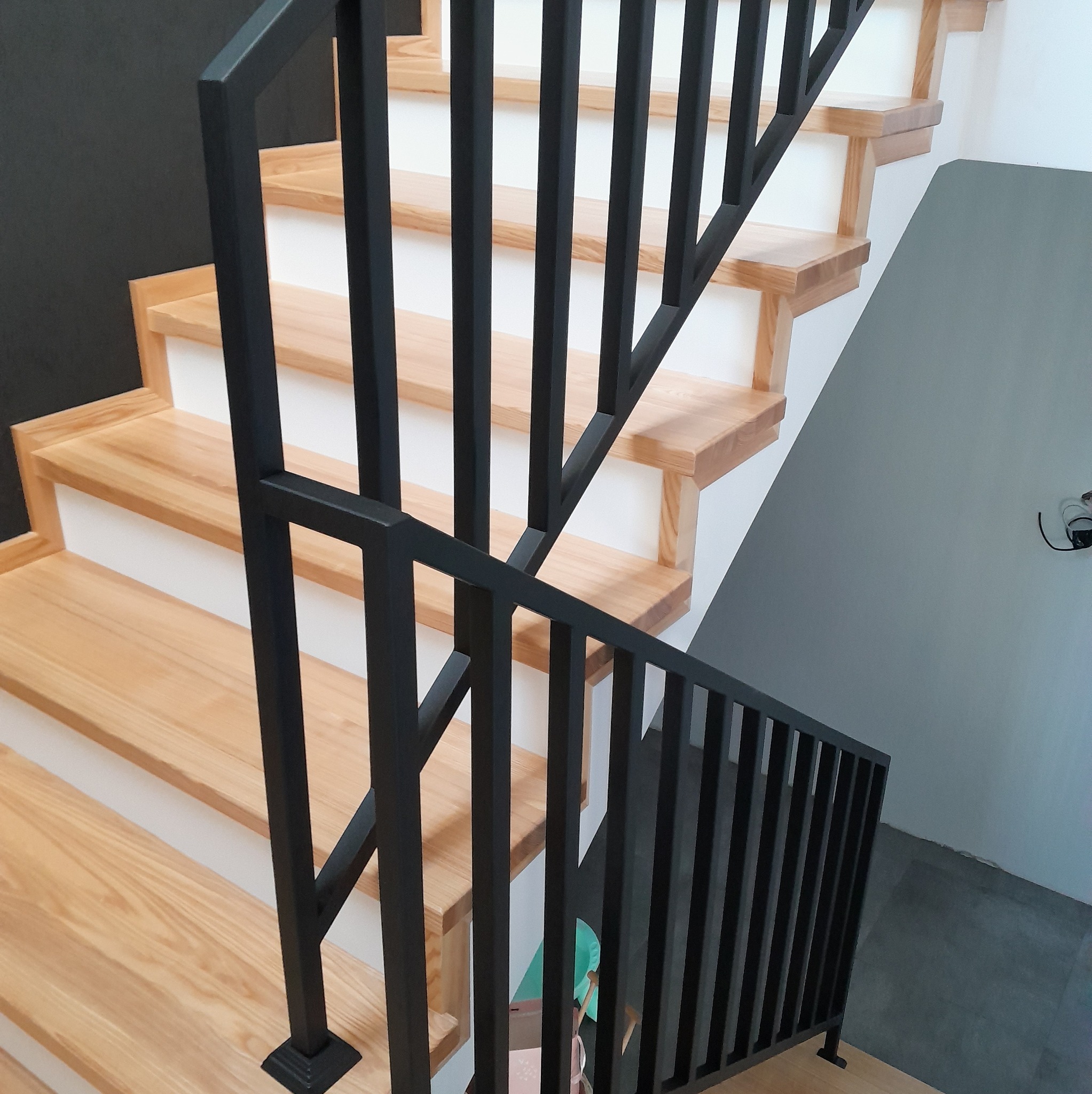 Nowoczesne schody z drewnianymi stopniami i czarną, metalową balustradą, widok z góry na dół, częściowo widoczna ściana w kolorze grafitowym.