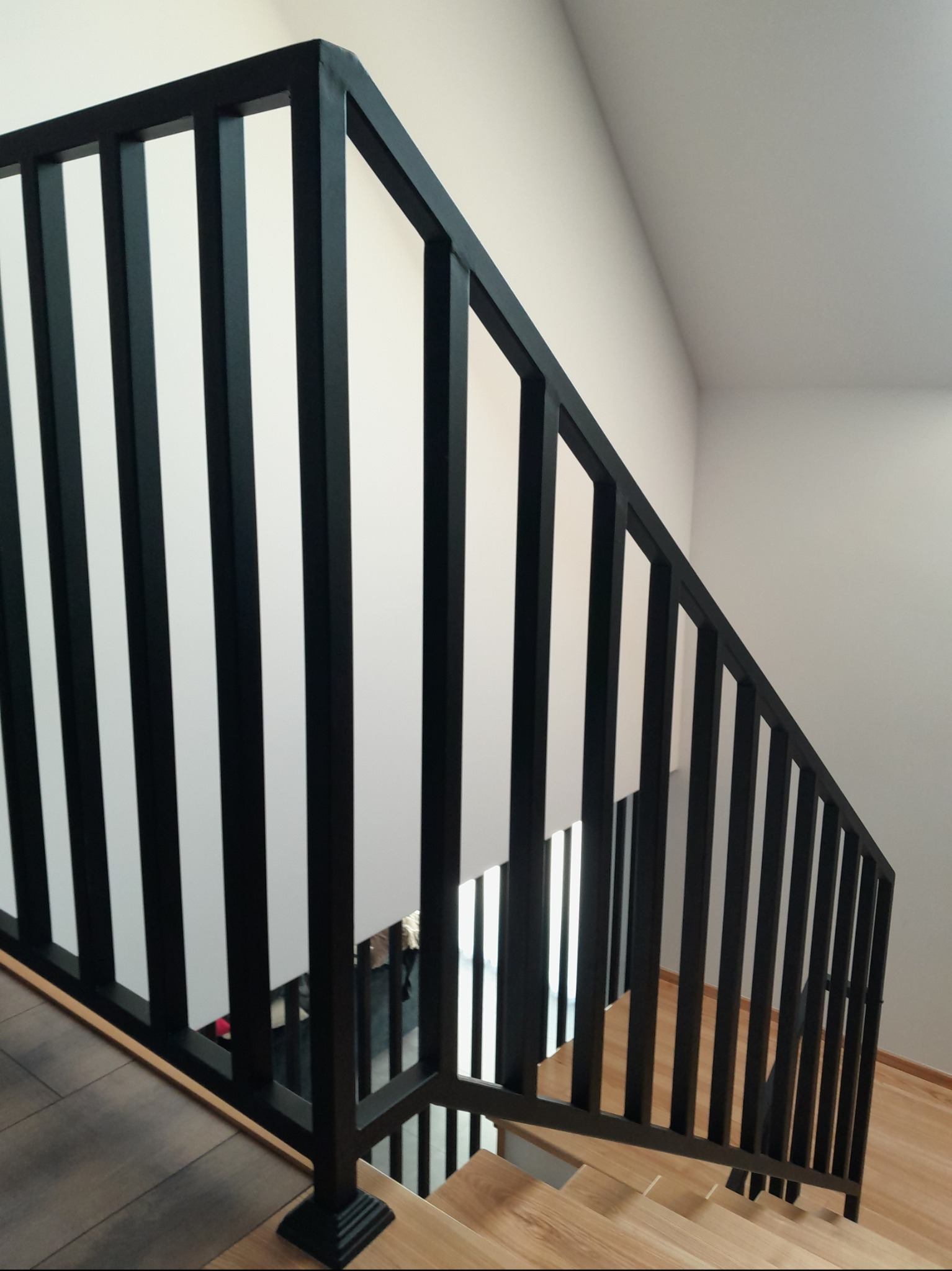 Minimalistyczne schody z drewnianymi stopniami i czarną, metalową balustradą z pionowymi szczeblami. Balustrada zakończona kwadratową podstawą. Widok od dołu schodów.