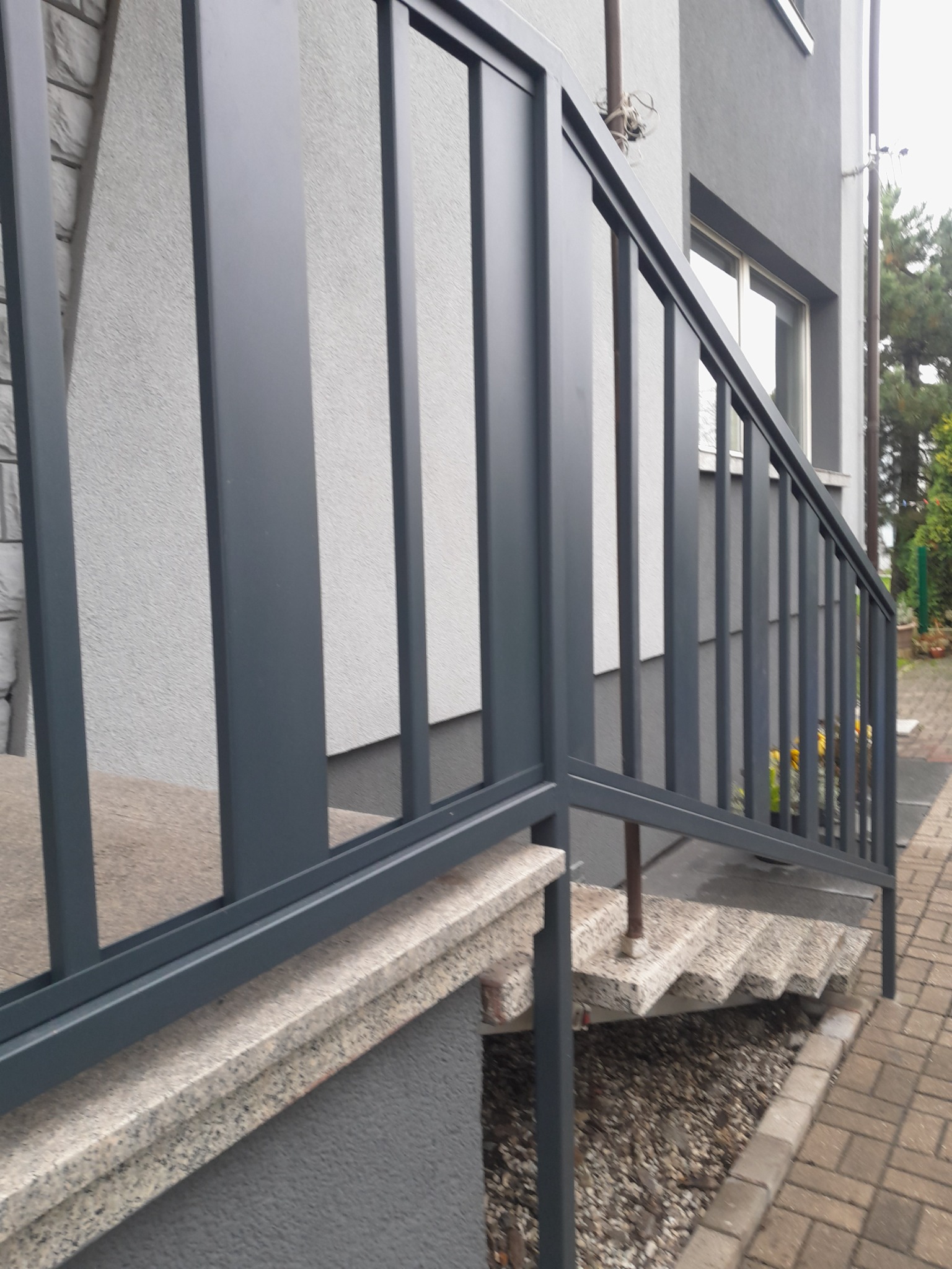 Szare metalowe barierki schodowe z prostymi pionowymi szczeblami, zamontowane na kamiennych schodach zewnętrznych prowadzących do domu z szarą elewacją.