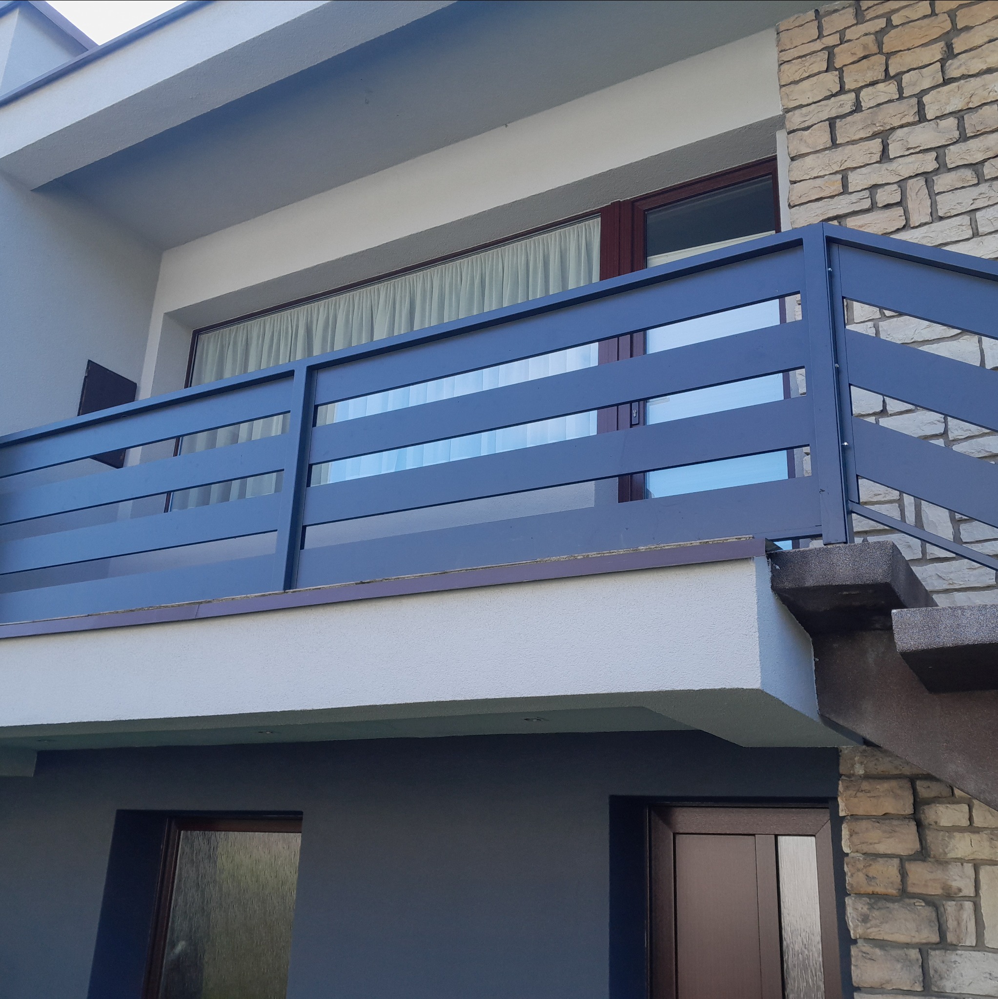 Nowoczesna balustrada balkonowa z metalu w kolorze grafitowym, z poziomymi panelami, zamontowana na balkonie budynku z elewacją z kamienia i tynkiem.