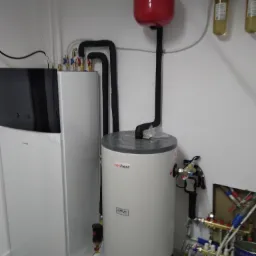 Instalacja pompy ciepła Daikin z zasobnikiem ciepłej wody, widoczne rury, zawory, manometr na czerwonym zbiorniku wyrównawczym, oraz filtry wody na ścianie.