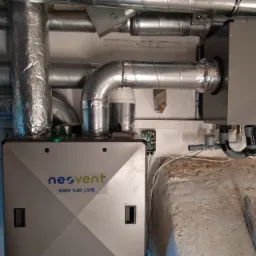 Szara centrala wentylacyjna Neovent KNV 520 LUX z podłączonymi przewodami wentylacyjnymi, widoczna instalacja elektryczna i fragment surowej ściany.