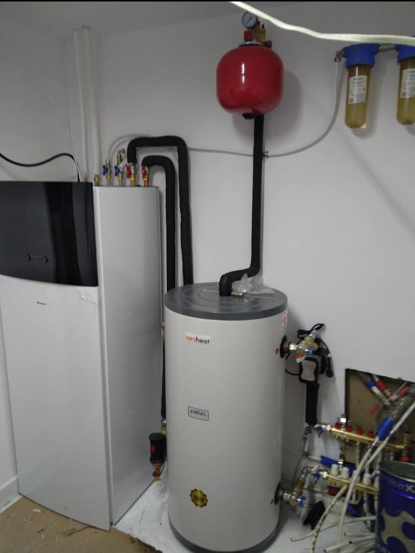 Instalacja grzewcza w kotłowni: biała pompa ciepła Daikin, szary zasobnik ciepłej wody z czerwonym naczyniem wzbiorczym, rury w izolacji. Elementy instalacji hydraulicznej widoczne w tle.