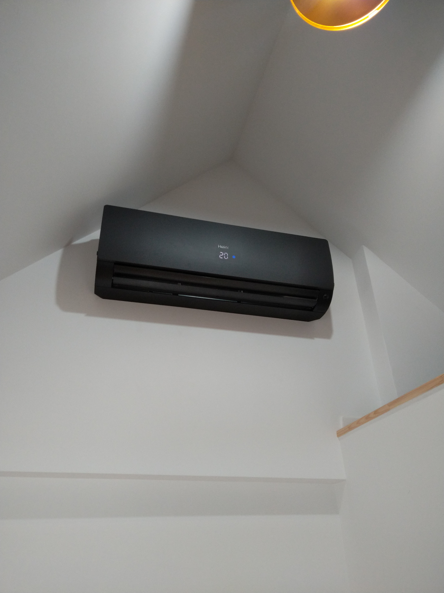 Czarny klimatyzator Haier zamontowany na białej ścianie w pokoju ze skosami, widoczny fragment oświetlenia sufitowego.