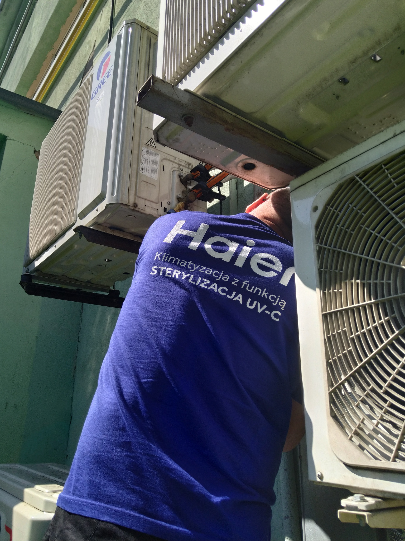 Serwisant w niebieskiej koszulce Haier naprawia klimatyzator zewnętrzny Gree, widoczne przewody i elementy montażowe. Drugi klimatyzator po prawej stronie.
