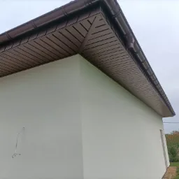 Narożnik budynku z jasną elewacją i brązowym podbitką dachową z widocznymi rynnami, widok z dołu na pochmurne niebo.