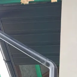 Fragment dachu z blachy trapezowej w kolorze grafitowym, widoczna rynna z tworzywa sztucznego oraz elementy drewnianej konstrukcji pomalowane na zielono, ujęcie z dołu.