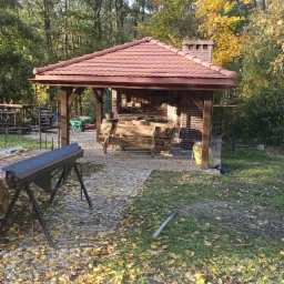 Zadaszona altana z ceglanym grillem i kominem, pokryta czerwoną dachówką, otoczona jesiennym lasem i brukowanym podłożem usłanym opadłymi liśćmi, z widocznym stołem warsztatowym.
