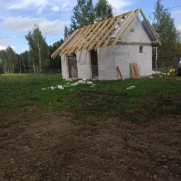 Niedokończony budynek z białych bloczków z drewnianą konstrukcją dachu, widoczne deski na ziemi, trawa i drzewa w tle, pochmurne niebo.