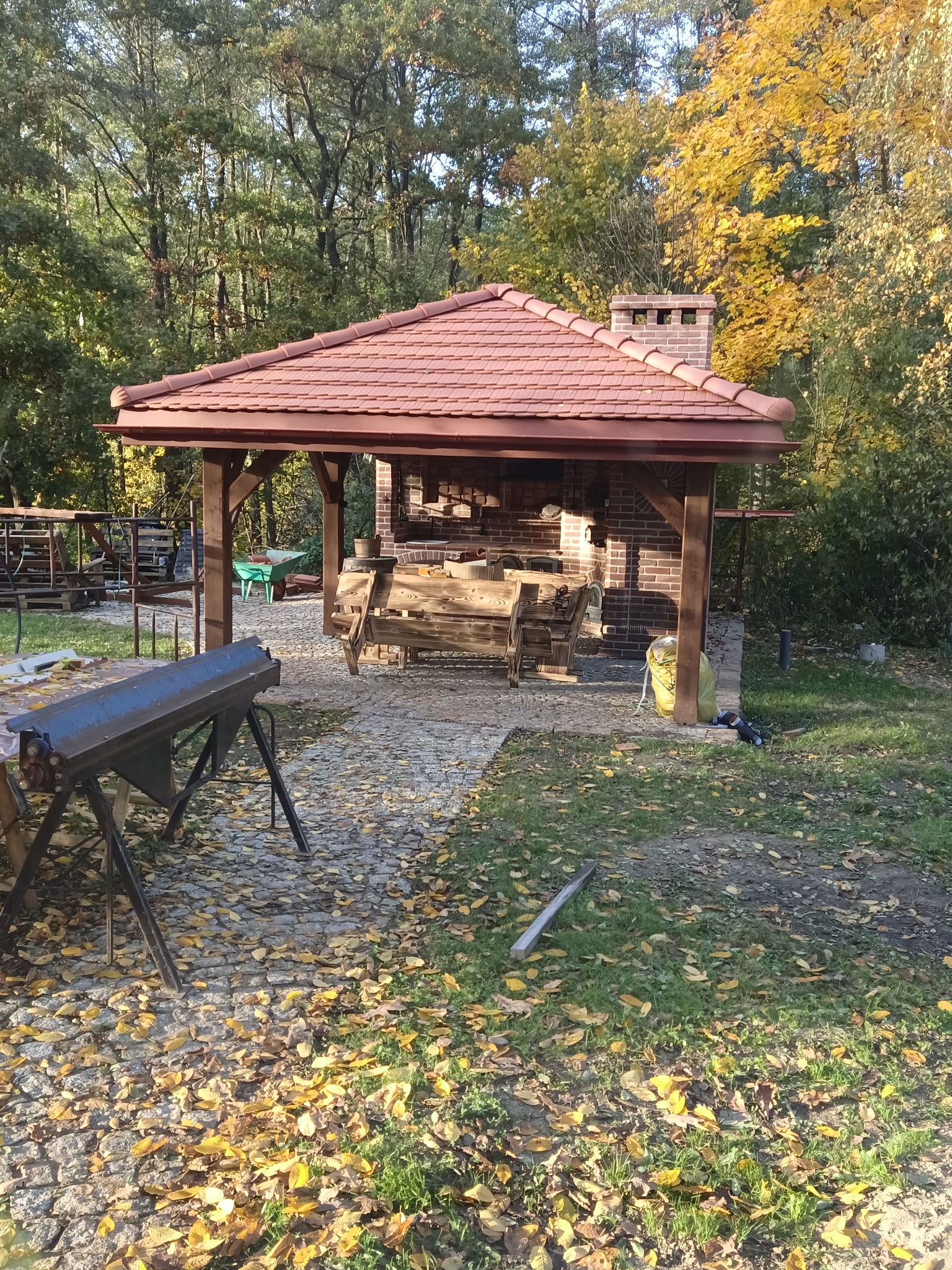 Zadaszona altana z ceglanym grillem i kominem, pokryta czerwoną dachówką, otoczona jesiennym lasem i brukowanym podłożem usłanym opadłymi liśćmi, z widocznym stołem warsztatowym.