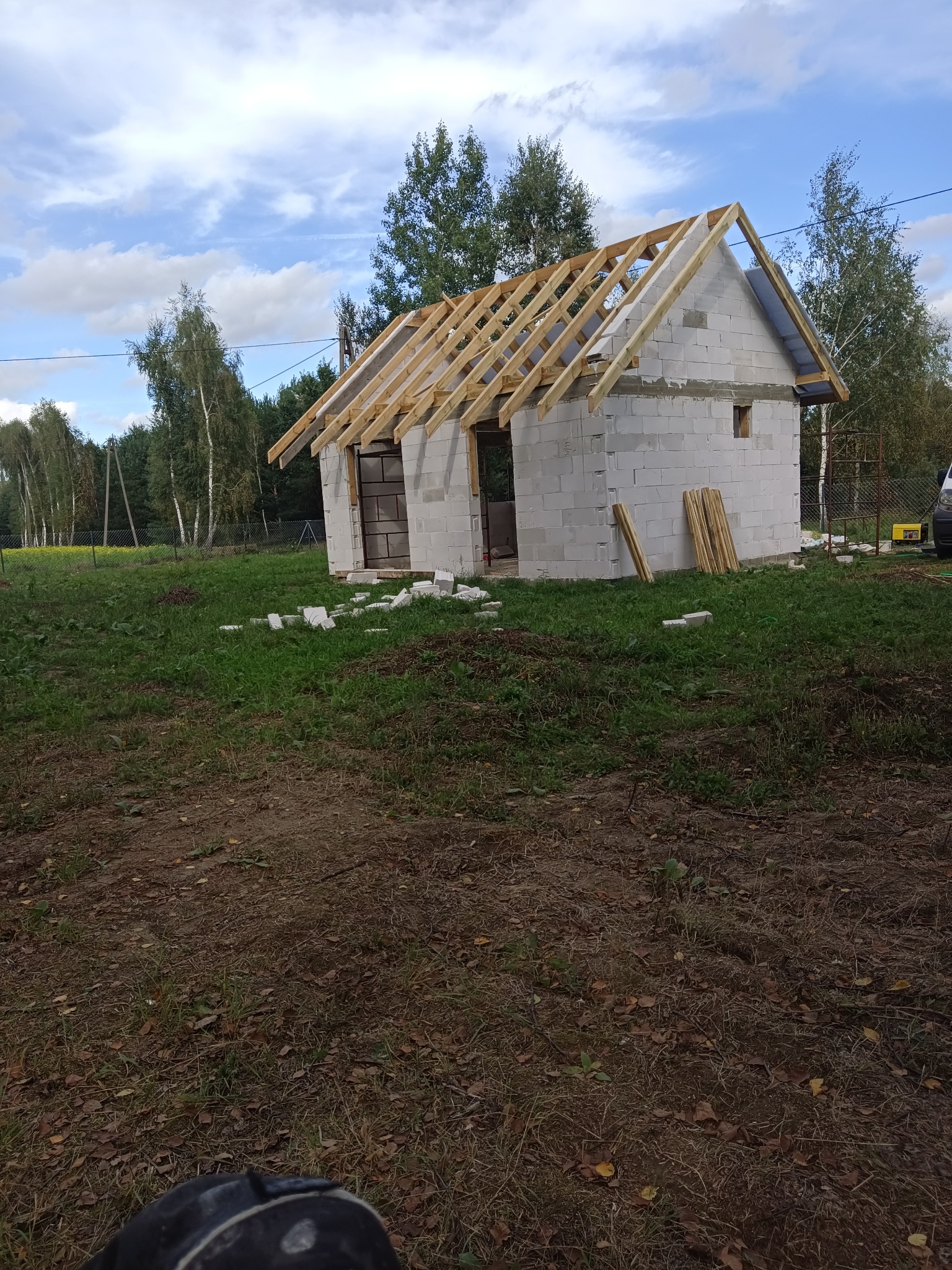 Niedokończony budynek z białych bloczków z drewnianą konstrukcją dachu, widoczne deski na ziemi, trawa i drzewa w tle, pochmurne niebo.