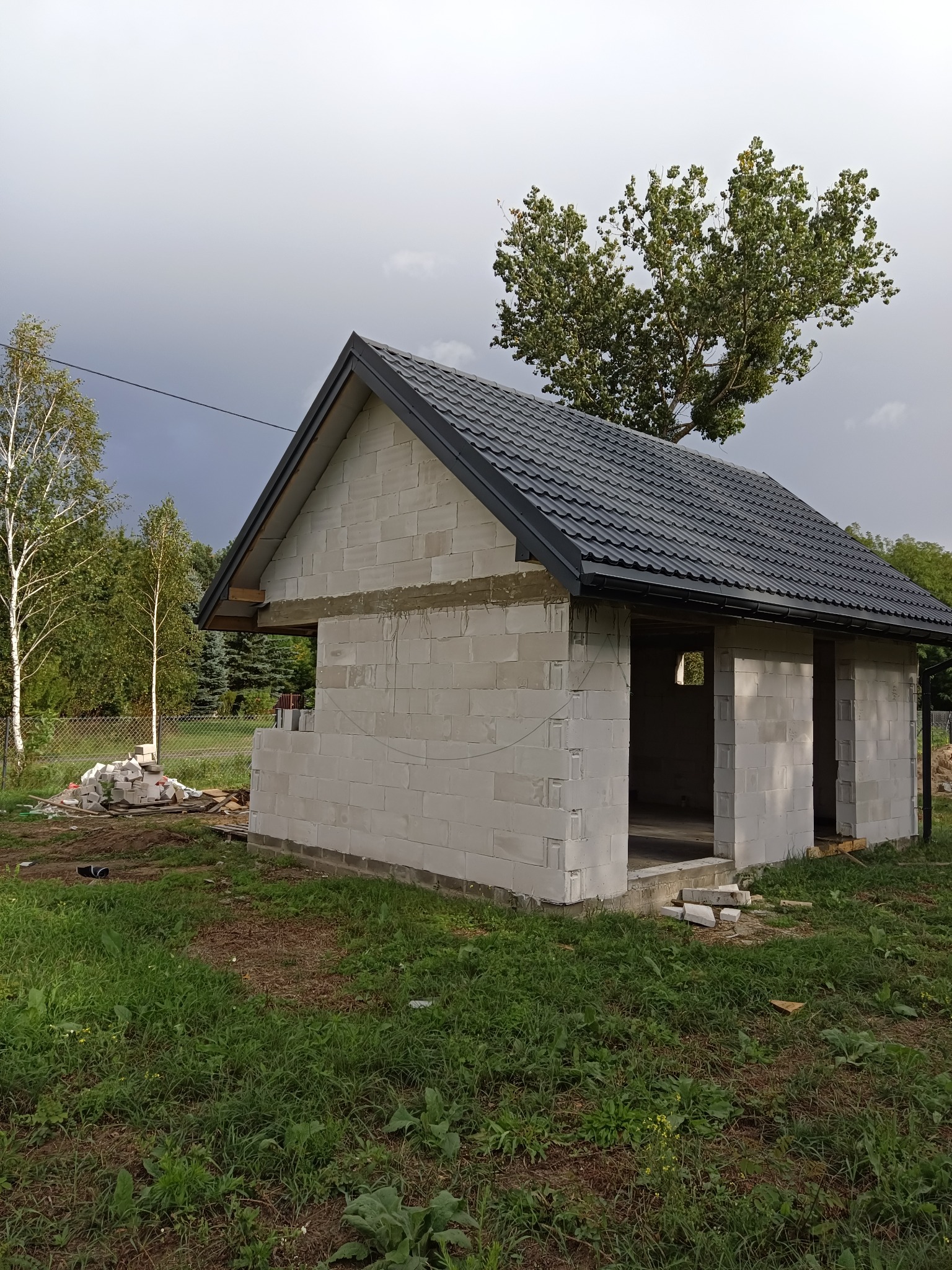Surowy budynek z białych bloczków z ukończonym, ciemnoszarym dachem z blachy, widok z zewnątrz na tle pochmurnego nieba i drzew