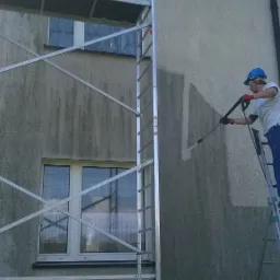 Pracownik w niebieskim kasku i kombinezonie, stojąc na drabinie, czyści elewację budynku myjką ciśnieniową; widoczny fragment rusztowania i okna.
