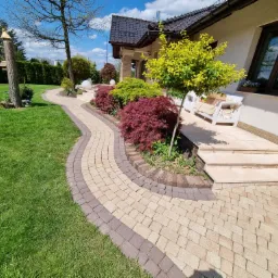 Kręta ścieżka z kostki brukowej w dwóch odcieniach prowadzi do domu z tarasem, otoczona zielonym trawnikiem i ozdobnymi krzewami.