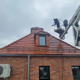 Pracownik na podnośniku koszowym myje ciśnieniowo dach z czerwonej dachówki, widoczna elewacja budynku z cegły i klimatyzator.