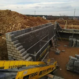 Wznoszenie muru oporowego z szarego bloczka betonowego na fundamencie, z widocznym rusztowaniem i żółtymi barierami ochronnymi TASSU, na tle wzgórza z ziemią i budynkami w oddali.