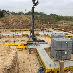 Wylewanie betonu do szalunków fundamentowych z użyciem pompy do betonu; widoczne zbrojenia, bloczki betonowe i pracownik w czerwonej kurtce.