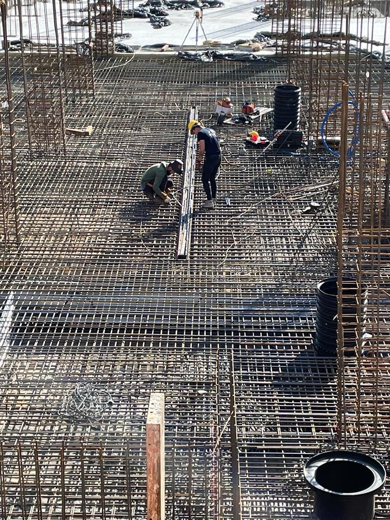 Dwóch zbrojarzy pracuje nad siatką zbrojeniową fundamentu, widoczne narzędzia i elementy konstrukcyjne, na dalszym planie niwelator.