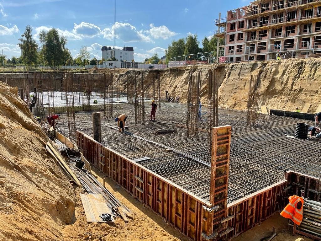 Budowa fundamentów: zbrojenie płyty fundamentowej z widocznymi szalunkami i pracującymi robotnikami na tle wykopu i budynku w stanie surowym otwartym.