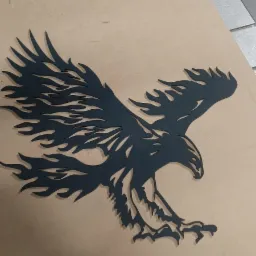 Wycięty z metalu lub tworzywa sztucznego, czarny, stylizowany orzeł z płomieniami zamiast piór na skrzydłach, leżący na płycie MDF z widocznymi śladami po frezowaniu.