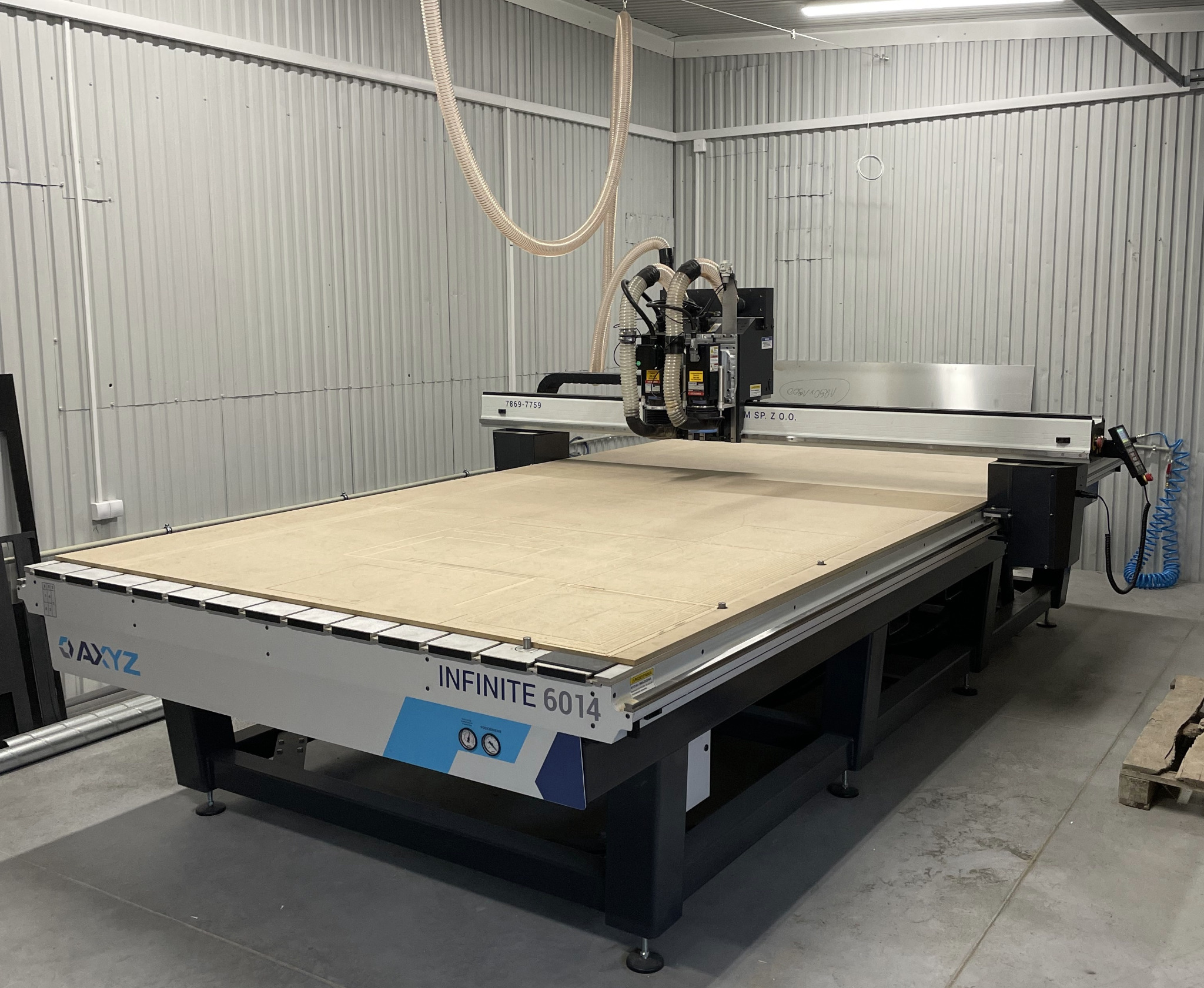 Duży ploter frezujący CNC marki AXYZ Infinite 6014 z blatem roboczym z płyty MDF, widoczny w hali produkcyjnej o ścianach z blachy trapezowej.