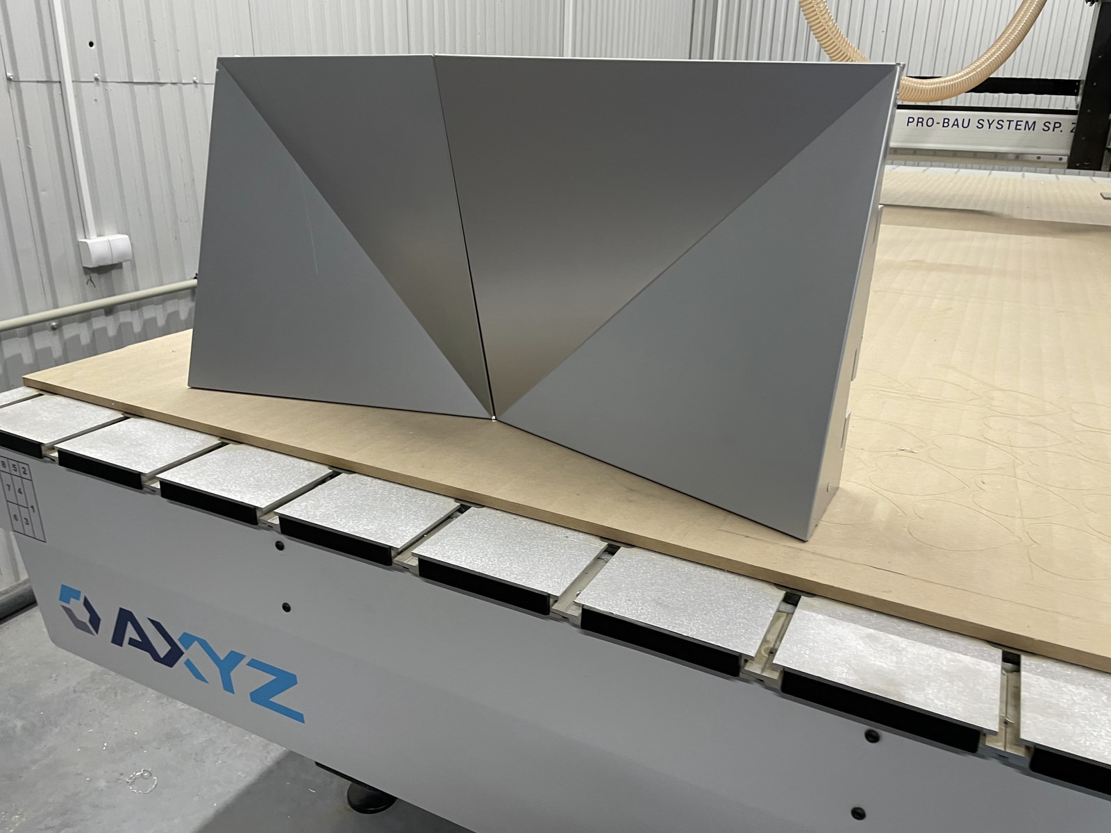 Szary, geometryczny element dekoracyjny z metalu, złożony z dwóch części, umieszczony na stole frezarki CNC AXYZ w warsztacie.