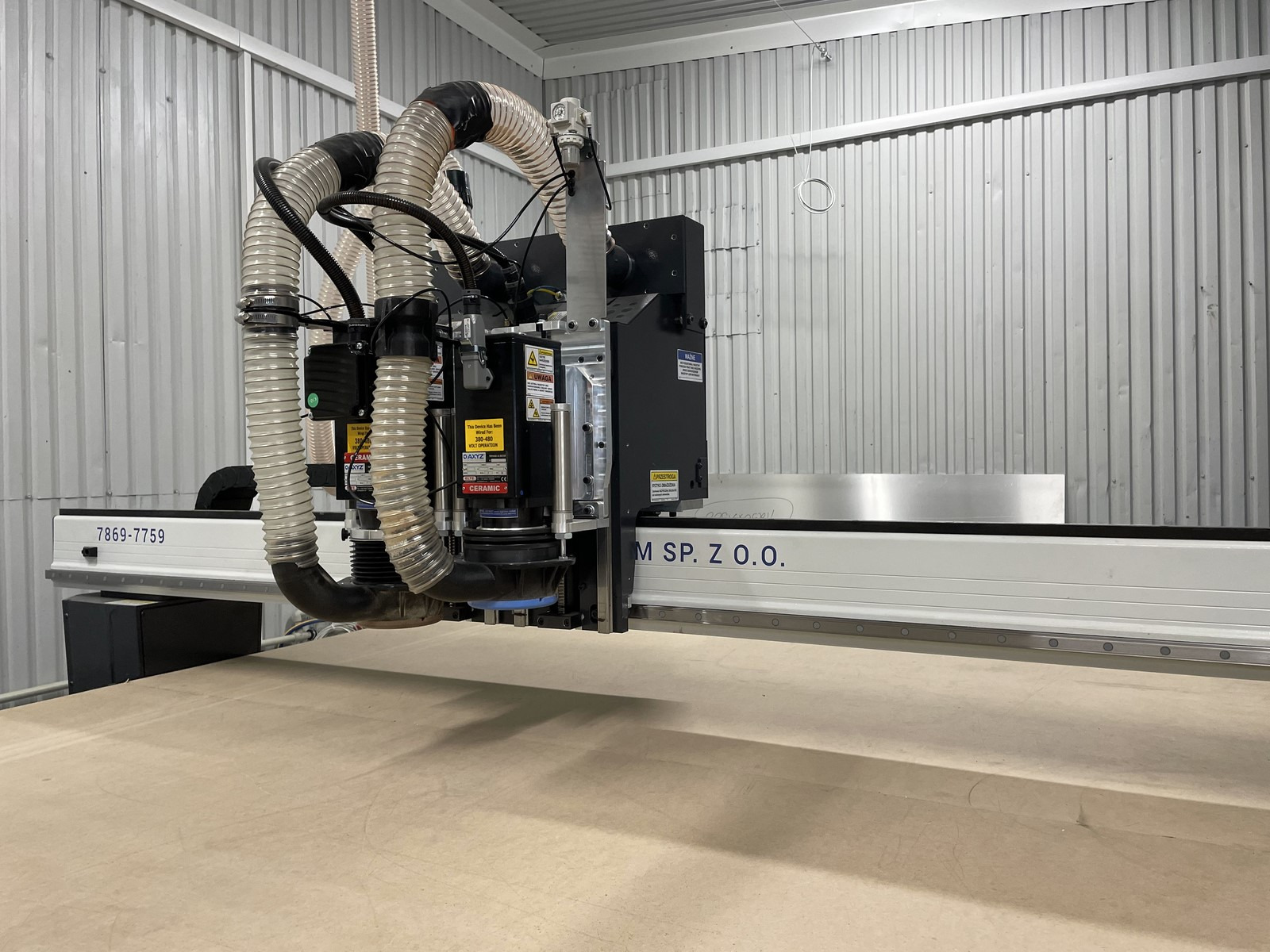 Duże urządzenie CNC z rurami odciągowymi i frezem, przygotowane do obróbki płyty z jasnego drewna, w hali o ścianach z blachy trapezowej.