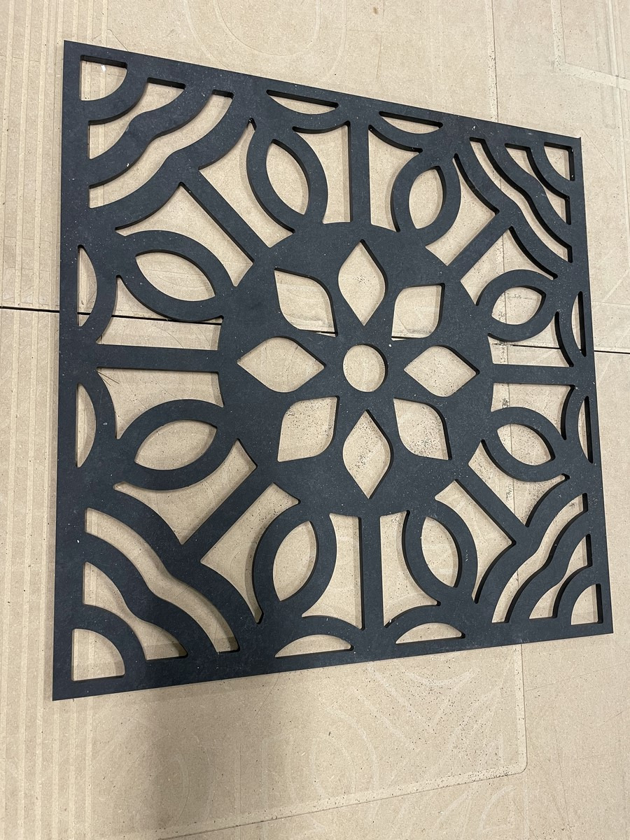 Czarny, ażurowy panel dekoracyjny z motywem kwiatowym i geometrycznym, wycięty z płyty na brązowym tle.