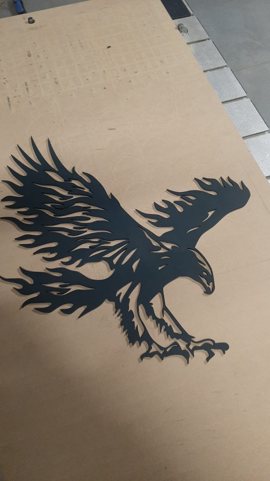 Wycięty z metalu lub tworzywa sztucznego, czarny, stylizowany orzeł z płomieniami zamiast piór na skrzydłach, leżący na płycie MDF z widocznymi śladami po frezowaniu.