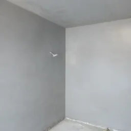 Surowe, szare wnętrze pokoju po nałożeniu tynku maszynowego; widoczne wystające przewody elektryczne i niedokończone wykończenie podłogi przy ścianie.