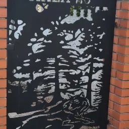 Czarna, metalowa brama z wycinanym laserowo motywem wilka w lesie i adresem 'MAŁA 26', osadzona w ceglanym murze.