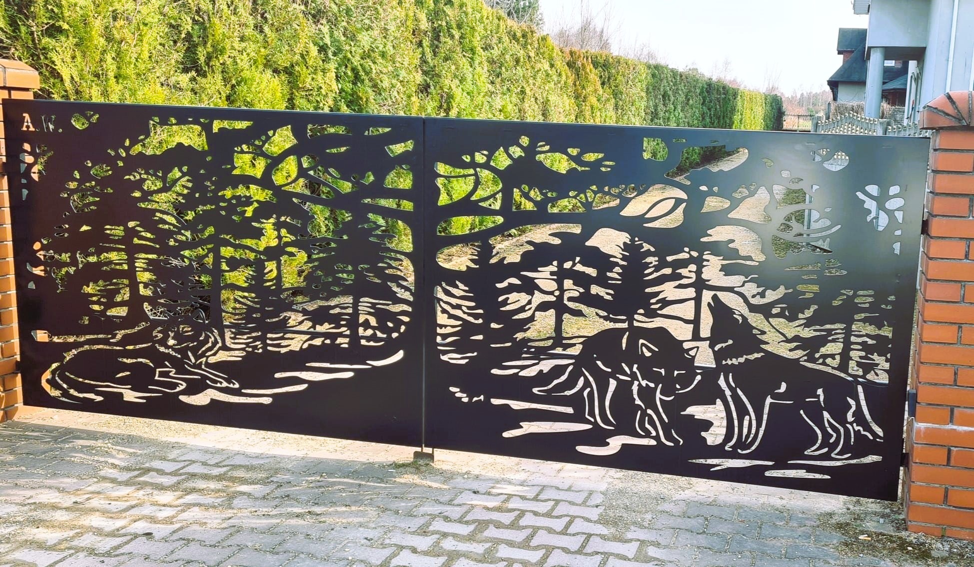 Metalowa brama wjazdowa z laserowo wycinanym motywem leśnym i wizerunkami wilków, osadzona między ceglanymi słupkami.