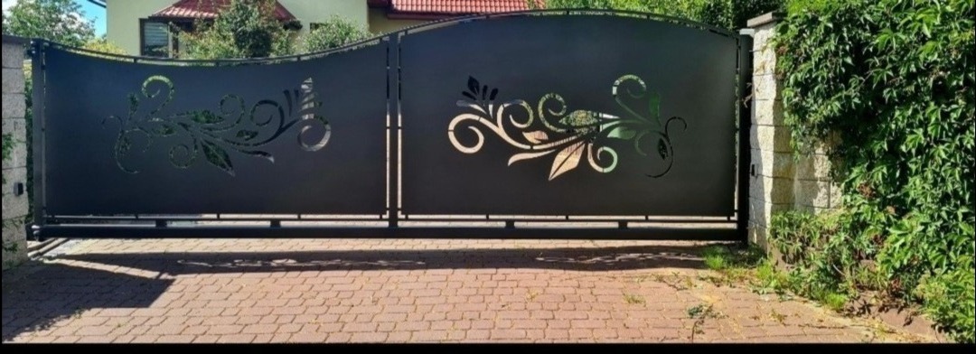 Czarna, metalowa brama wjazdowa z laserowo wycinanymi, ażurowymi wzorami roślinnymi, zamontowana pomiędzy kamiennymi słupkami porośniętymi bluszczem, na tle domu z czerwoną dachówką.