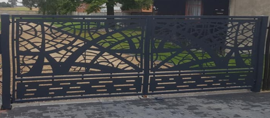 Metalowa brama wjazdowa z ażurowym, nowoczesnym wzorem na tle zieleni ogrodu, wykonana z ciemnoszarego metalu.
